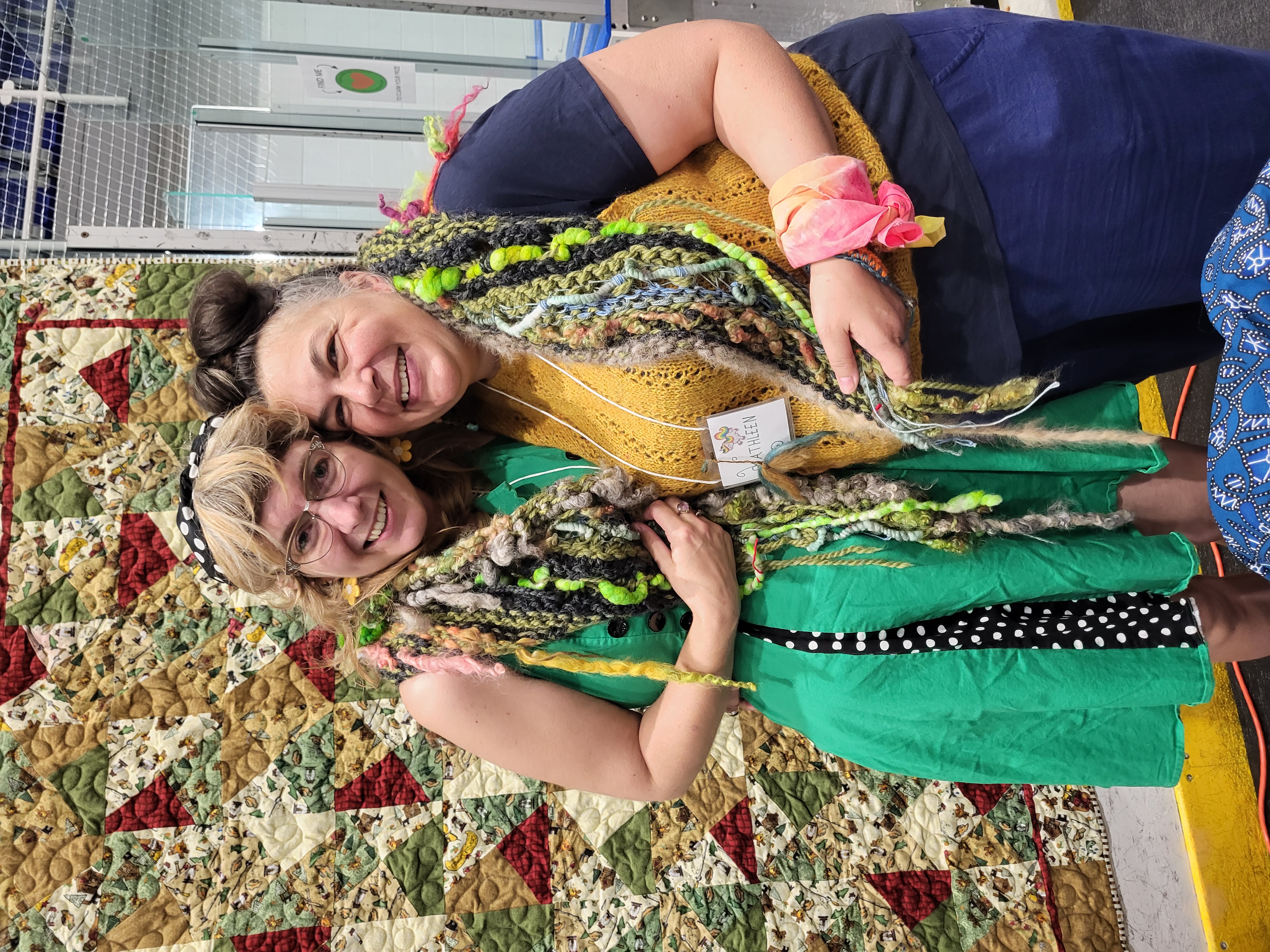 Two women huddle under a knitted shawl
